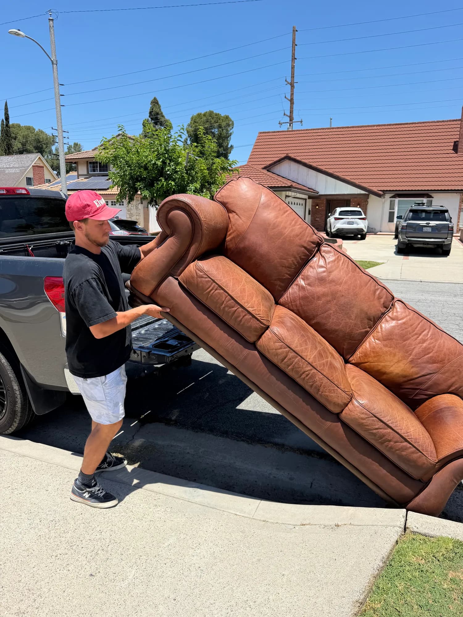 A man tipping a brown leather sofa into the bed of a pickup truck at the curb outside a house