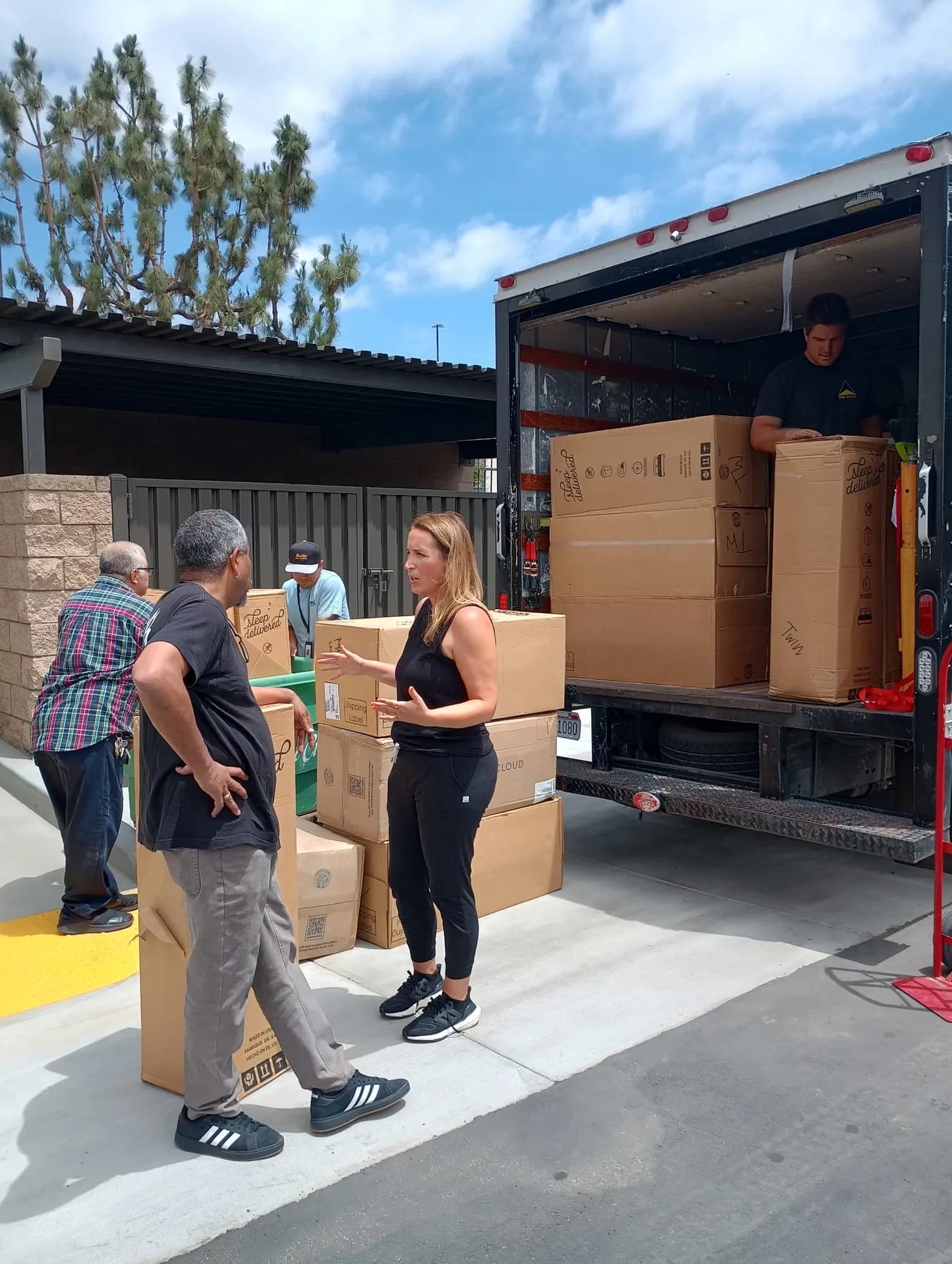 A box truck backed up outside a building with mattress cartons stacked inside, and a woman in black talking with two men beside more boxes on the pavement